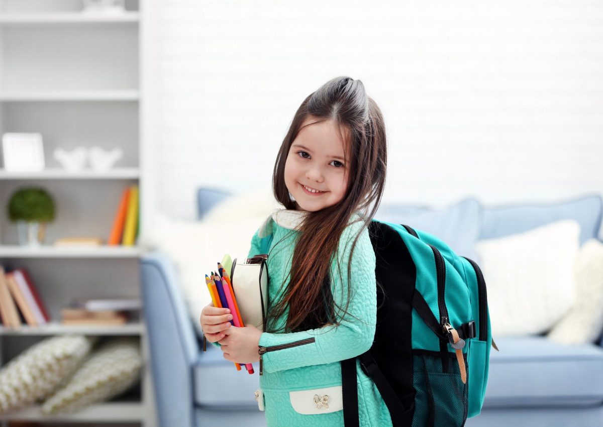 Little,Girl,With,Green,Back,Pack,Holding,Stationery,In,Living